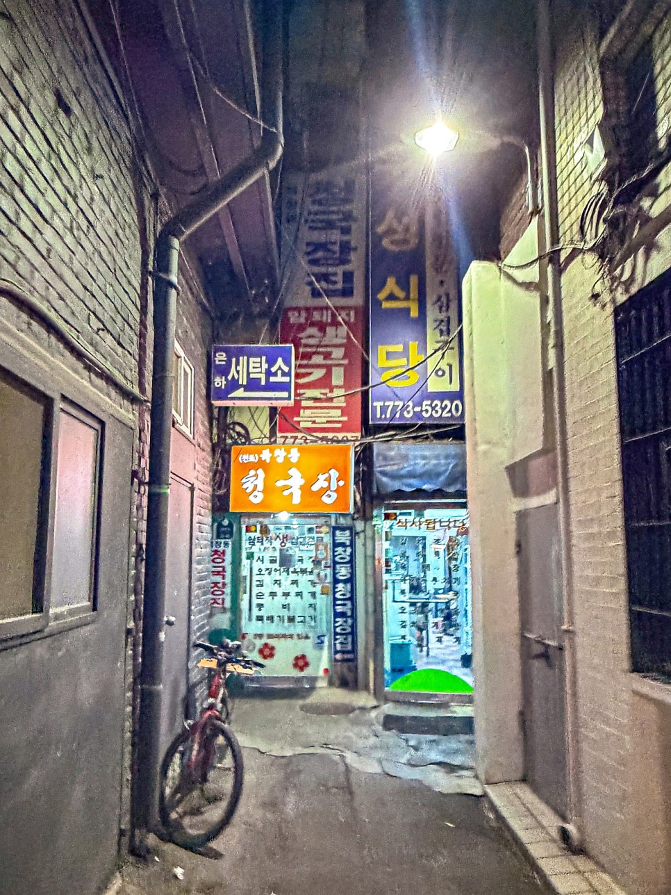 밤 골목 안쪽에 전화번호 T.773-5320이 적힌 북창동 우성식당 세로 간판과 청국장집 간판이 켜져 있고 자전거가 세워져 있다
